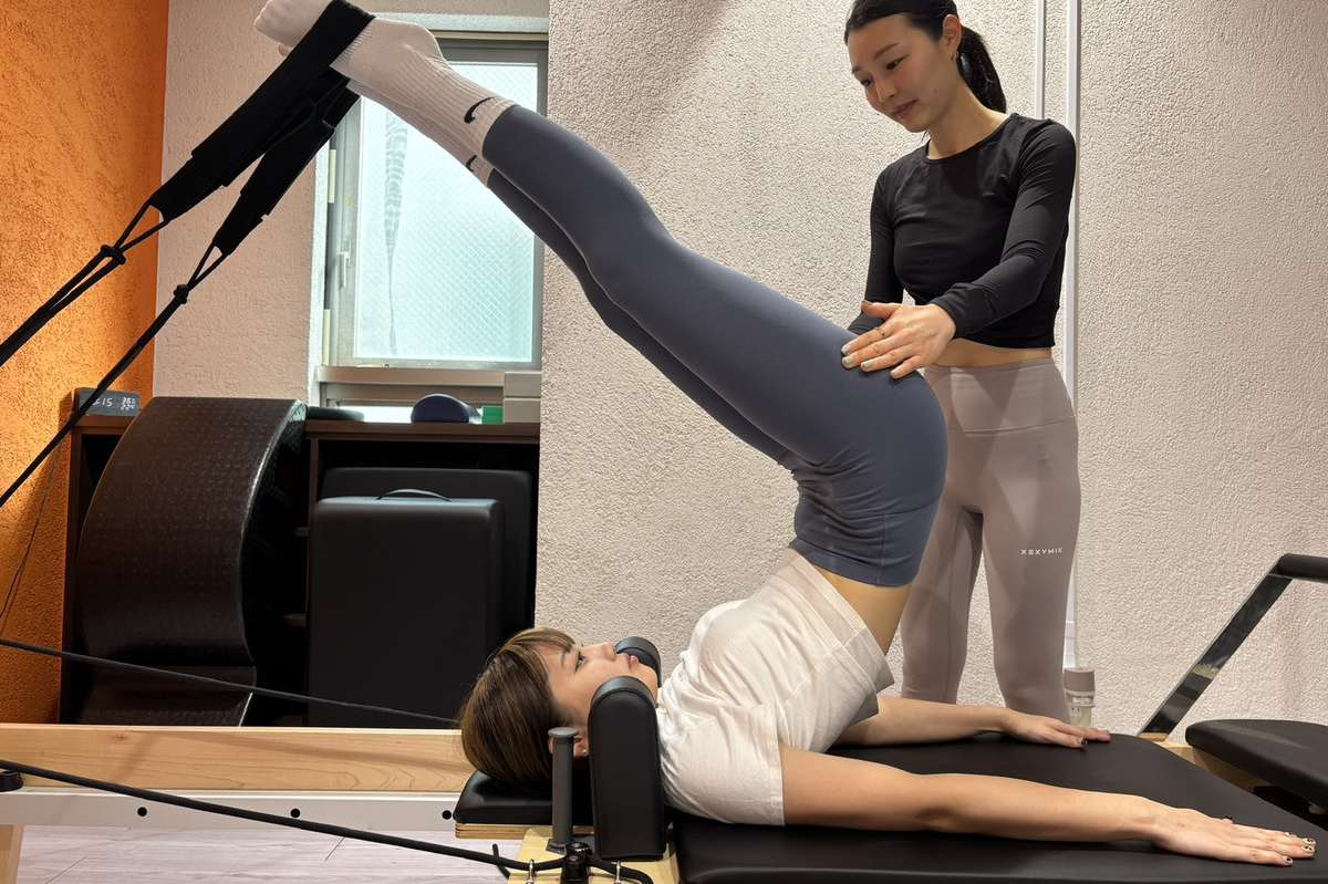 リフォーマーに仰向けになり、ストラップに足をかけて腰を持ち上げる種目を、トレーナーがそばで確認する様子