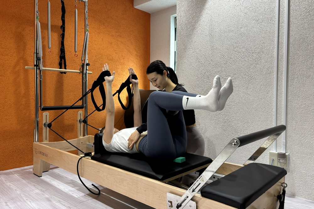 リフォーマーに仰向けになり、ストラップに足をかけて脚を動かす種目を、トレーナーが横で確認する様子